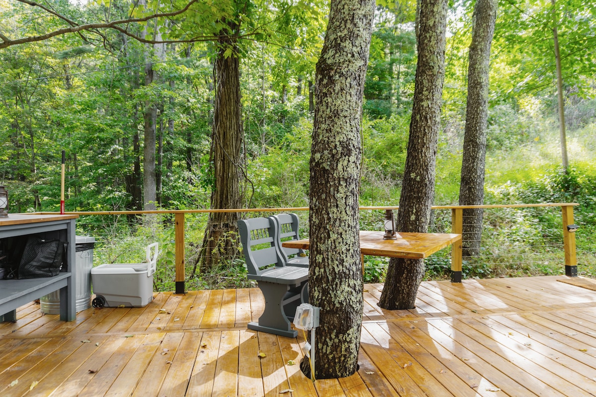 A spacious wooden deck is surrounded by tall trees, offering a serene outdoor space. A round table with two chairs sits at one end, while a grill station is nearby, inviting guests to enjoy al fresco dining amidst the natural setting.