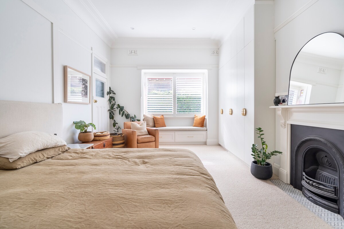 A spacious bedroom features a large bed with a soft, neutral-colored duvet. A cozy seating area is visible, consisting of two orange chairs and a low wooden table. Natural light enters through a window, and a decorative fireplace adds charm to the room.
