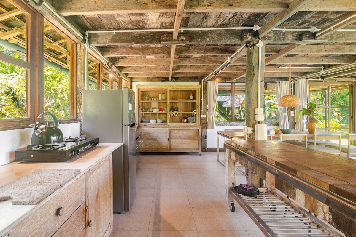 The open kitchen features rustic wooden cabinetry and a rustic wood island with a metal rack underneath. A modern refrigerator is positioned alongside a gas stove. Large windows invite natural light, showcasing green surroundings and creating a bright, airy atmosphere.