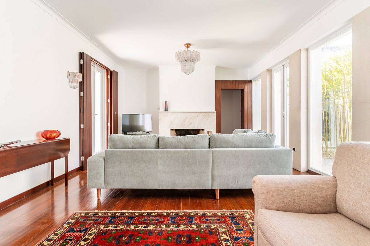 A spacious living room is presented, featuring a plush sofa and an armchair positioned on a vibrant area rug. Large windows allow natural light to fill the space, while a television and a decorative console table enhance the modern aesthetic.