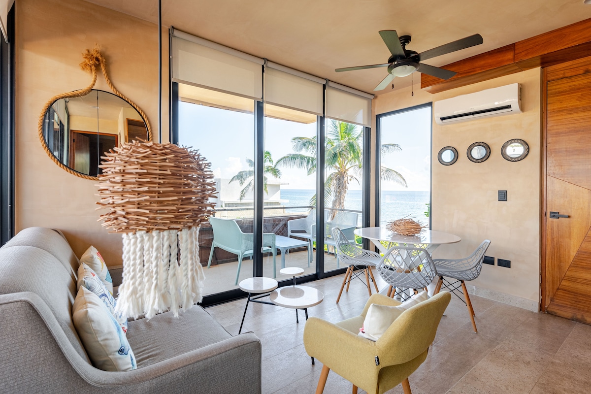 A cozy living area features large glass doors that provide a view of the ocean and palm trees outside. A comfortable sofa and an accent chair surround a circular dining table, while a unique light fixture hangs above. Air conditioning units and a large mirror enhance the space.