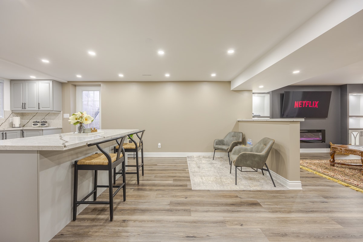 A contemporary living space is showcased, featuring a breakfast bar with sleek stools. Soft lighting illuminates the area, highlighting a minimalist design. A cozy seating arrangement is positioned nearby, with a large screen displaying 'NETFLIX' on the wall. Light wood flooring enhances the overall modern aesthetic.