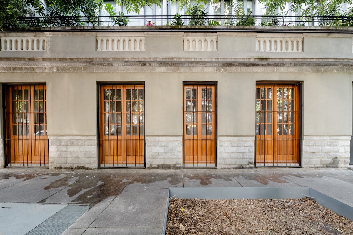 The exterior of the building features a light-colored facade with four wooden doors framed by metal grilles. A stone base supports the structure, and greenery from nearby trees enhances the atmosphere. A paved area is visible in front of the building.
