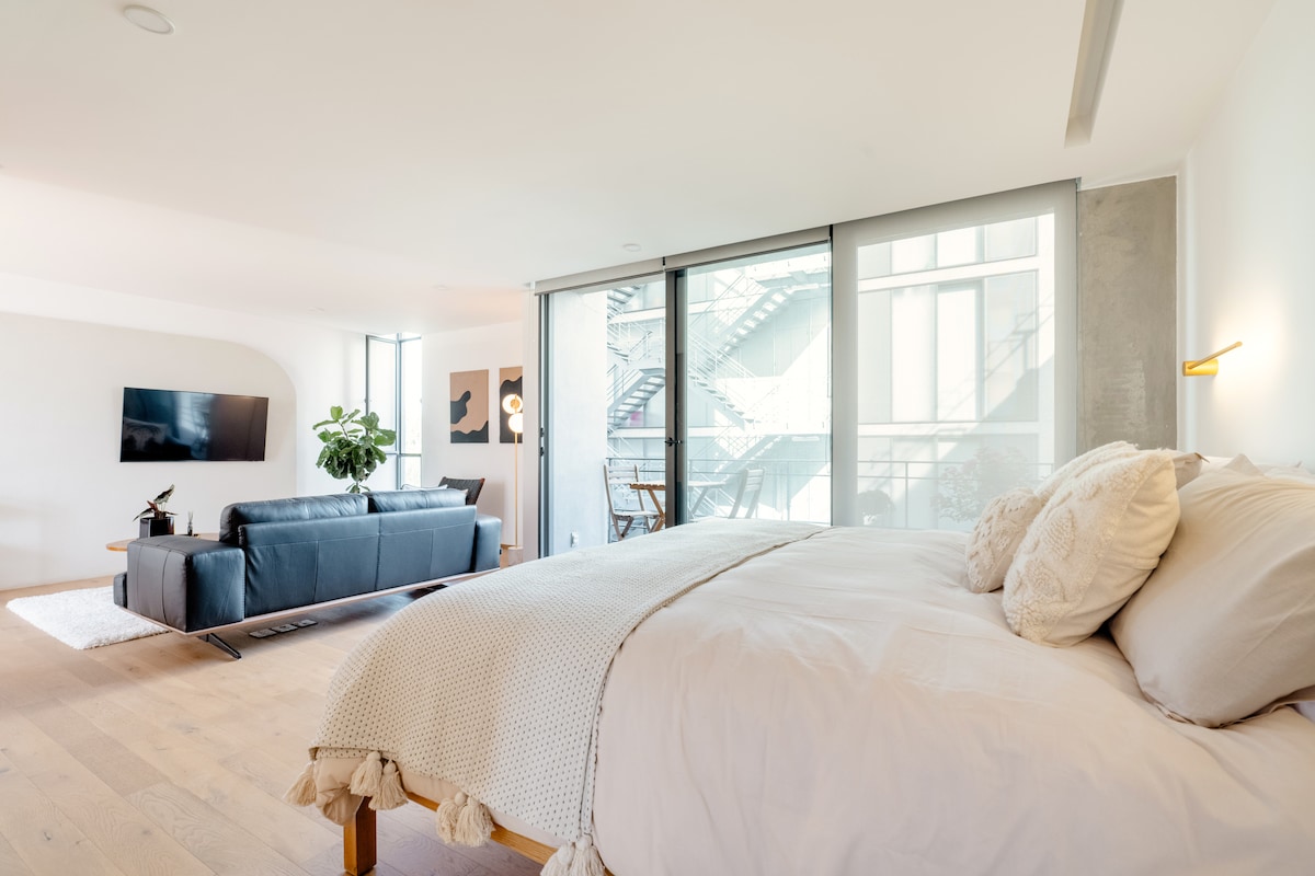 A spacious loft is presented, featuring a light-filled area with large windows. A comfortable bed is positioned in the foreground, while a sleek sofa and a minimalist television setup are seen in the living area. A touch of greenery from a potted plant adds a natural element.