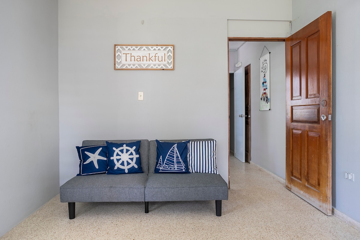 A simple living area features a gray sofa adorned with navy blue and white decorative pillows depicting nautical themes. A 'Thankful' sign mounted on the wall adds a personal touch, while a wooden door and an adjacent entryway can be seen in the background.