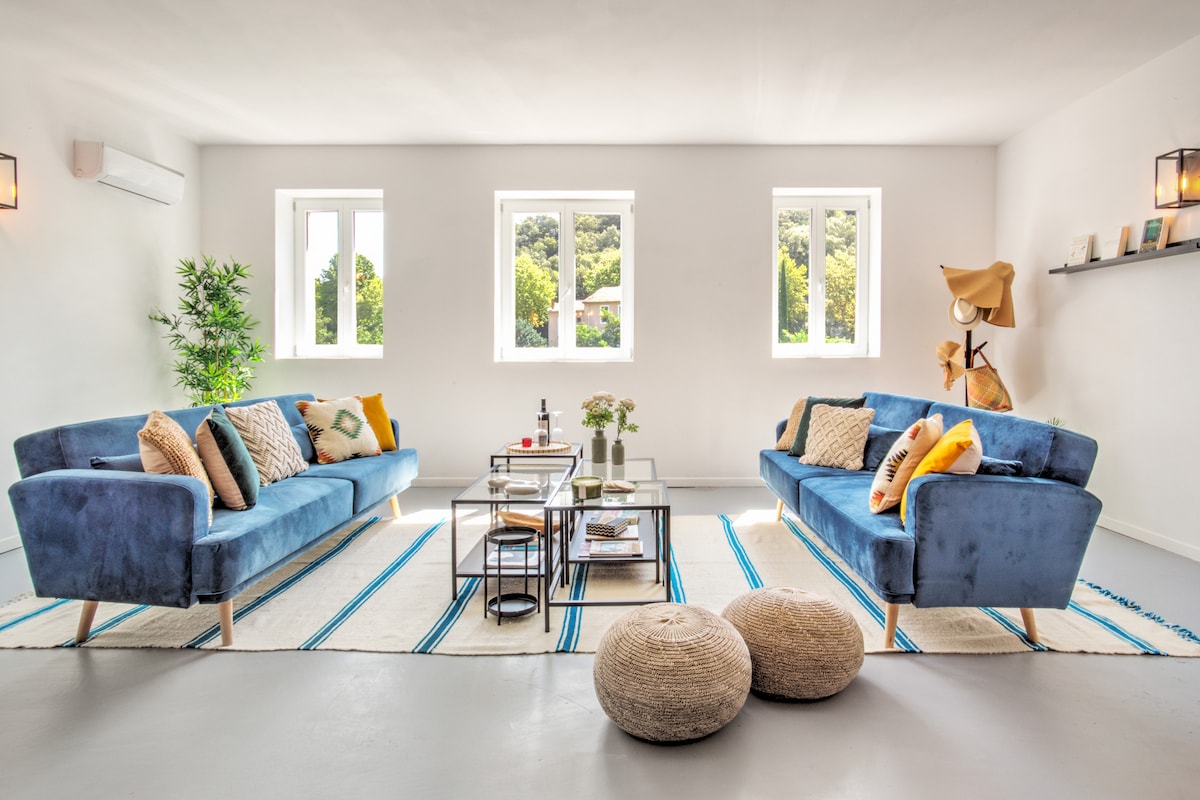 A spacious living area is presented with two blue sofas, adorned with colorful cushions. A glass coffee table sits centrally, complemented by decorative items. Natural light floods the room through two windows, while a textured area rug and woven poufs enhance the inviting environment.