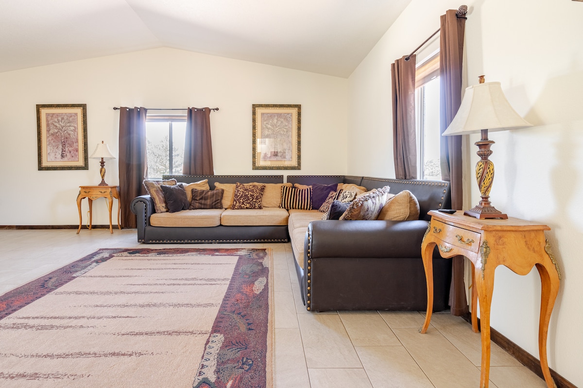 A spacious living area features a large sectional sofa adorned with decorative cushions. Natural light filters through windows draped with dark curtains, and a stylish side table stands nearby. Artwork is displayed on the walls, complementing the inviting atmosphere.