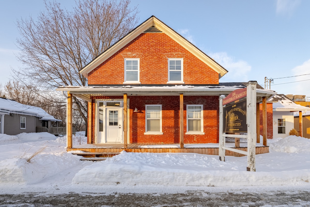 A classic two-story brick building is shown, featuring a welcoming porch and large front windows. Snow covers the ground and surrounds the entrance, while trees with bare branches contribute to the winter scenery. The sign indicates the building's purpose as a welcoming accommodation.