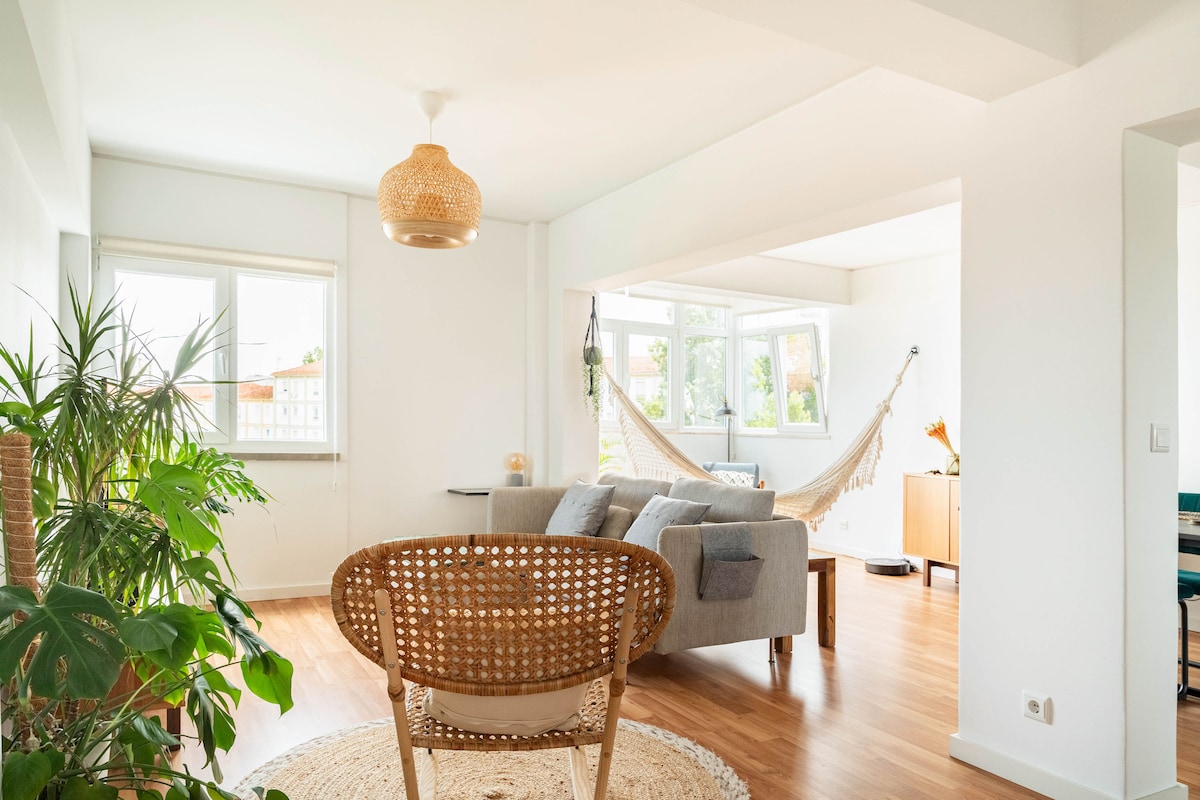 A bright living area is featured, with large windows allowing natural light. A comfortable gray sofa is positioned near a rattan chair, and a hammock is suspended in the corner. Lush green plants add a touch of nature to the space.