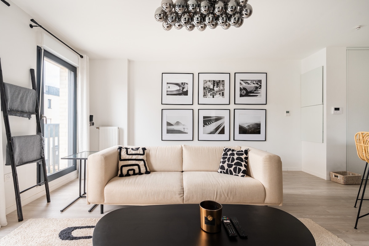 A bright living area features a light-colored sofa adorned with contrasting cushions. A sleek black coffee table sits in front, and a modern chandelier hangs above. Framed black-and-white photographs line the wall, complemented by a floor-to-ceiling window providing natural light.