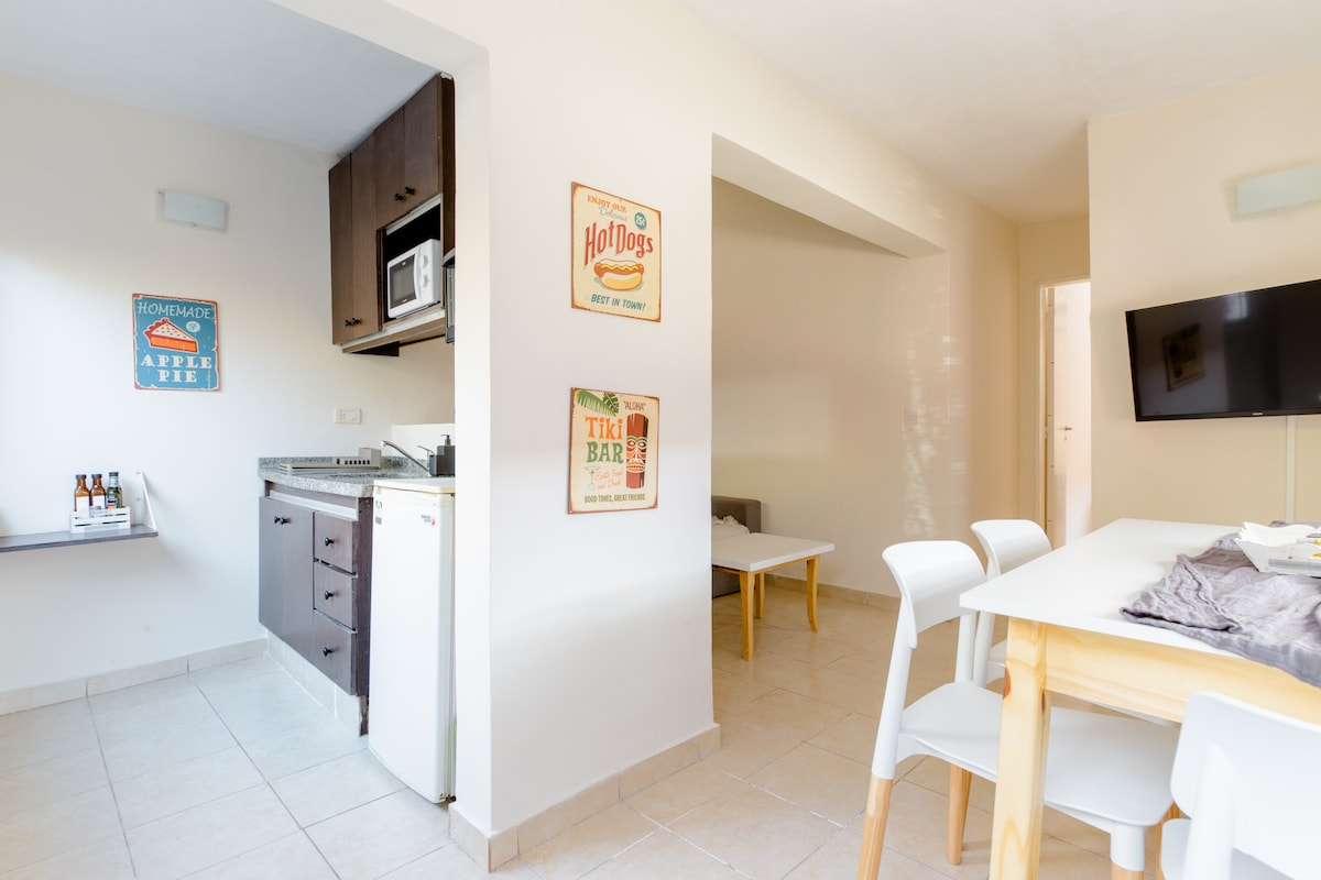 A bright and open living space features a kitchenette with modern appliances and light-colored cabinetry. A dining table with white chairs is positioned in front of a wall adorned with vintage posters. A TV is mounted on the opposite wall, enhancing the casual atmosphere.