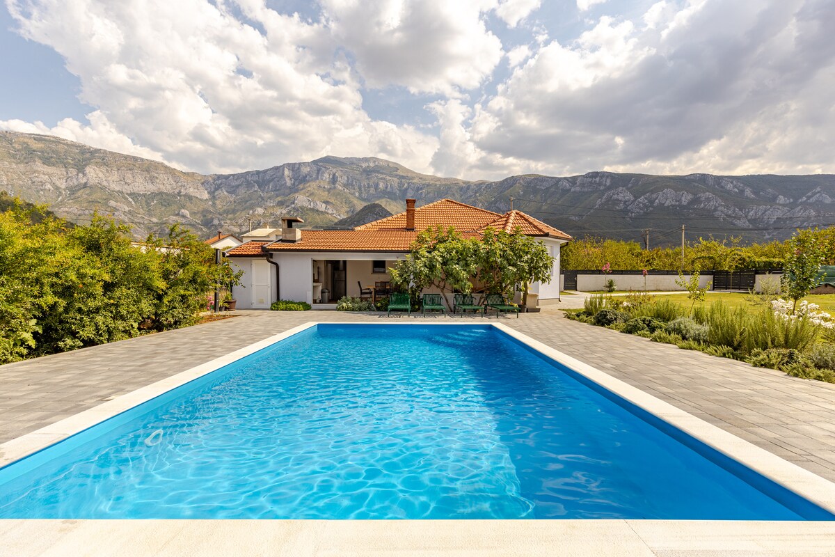A tranquil outdoor pool is surrounded by a spacious terrace, featuring a modern home in the background. Lush greenery frames the area, and majestic mountains rise in the distance under a partly cloudy sky, providing a picturesque setting for relaxation.