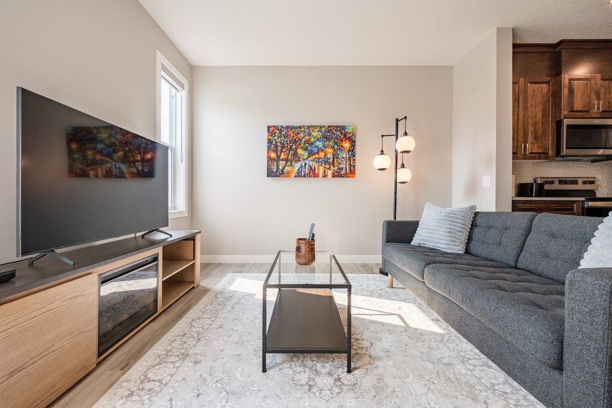 A modern living area is highlighted by a comfortable gray sofa facing a large flat-screen TV. A glass coffee table rests on a neutral area rug, complementing the light wood flooring. A colorful artwork adorns the wall, with a kitchen area visible in the background.