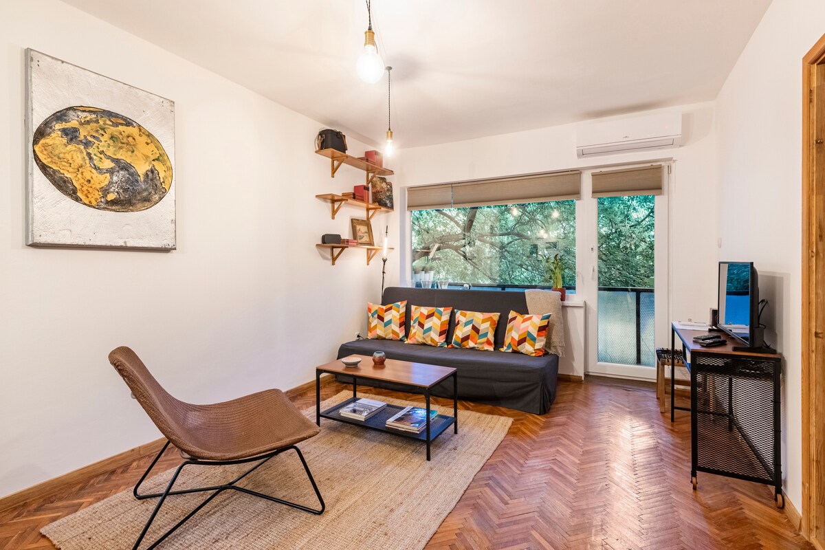 A modern living area features a comfortable sofa adorned with colorful cushions. A minimalist shelf displays decorative items. Natural light filters through glass doors, revealing a view of green trees outside. A small table and a television on a metal stand enhance the functional space.