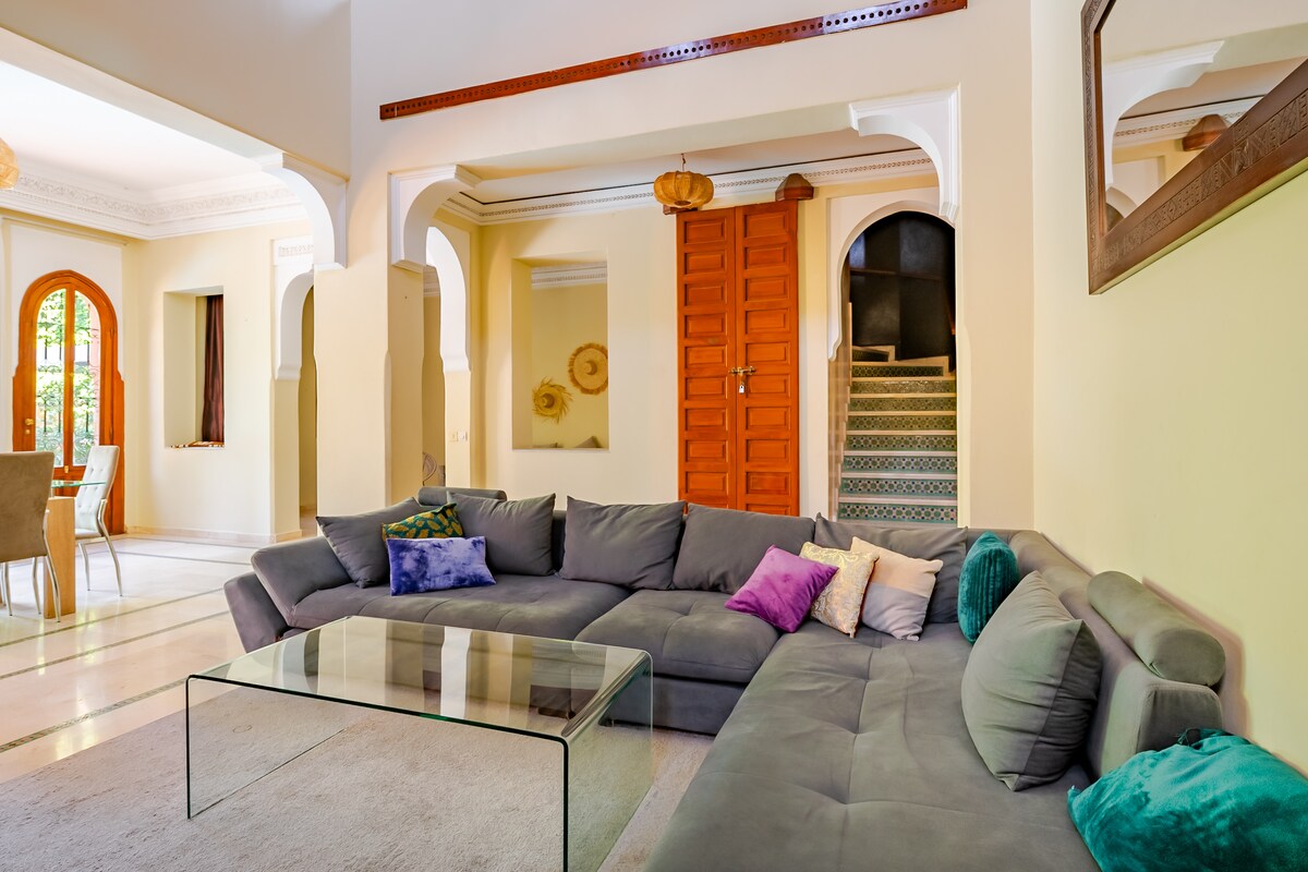 A spacious living area features a large, comfortable sectional sofa adorned with colorful pillows. A glass coffee table sits in front, complementing the neutral tones of the decor. Arched doorways create an open feel, leading towards a dining area and staircase in the background.