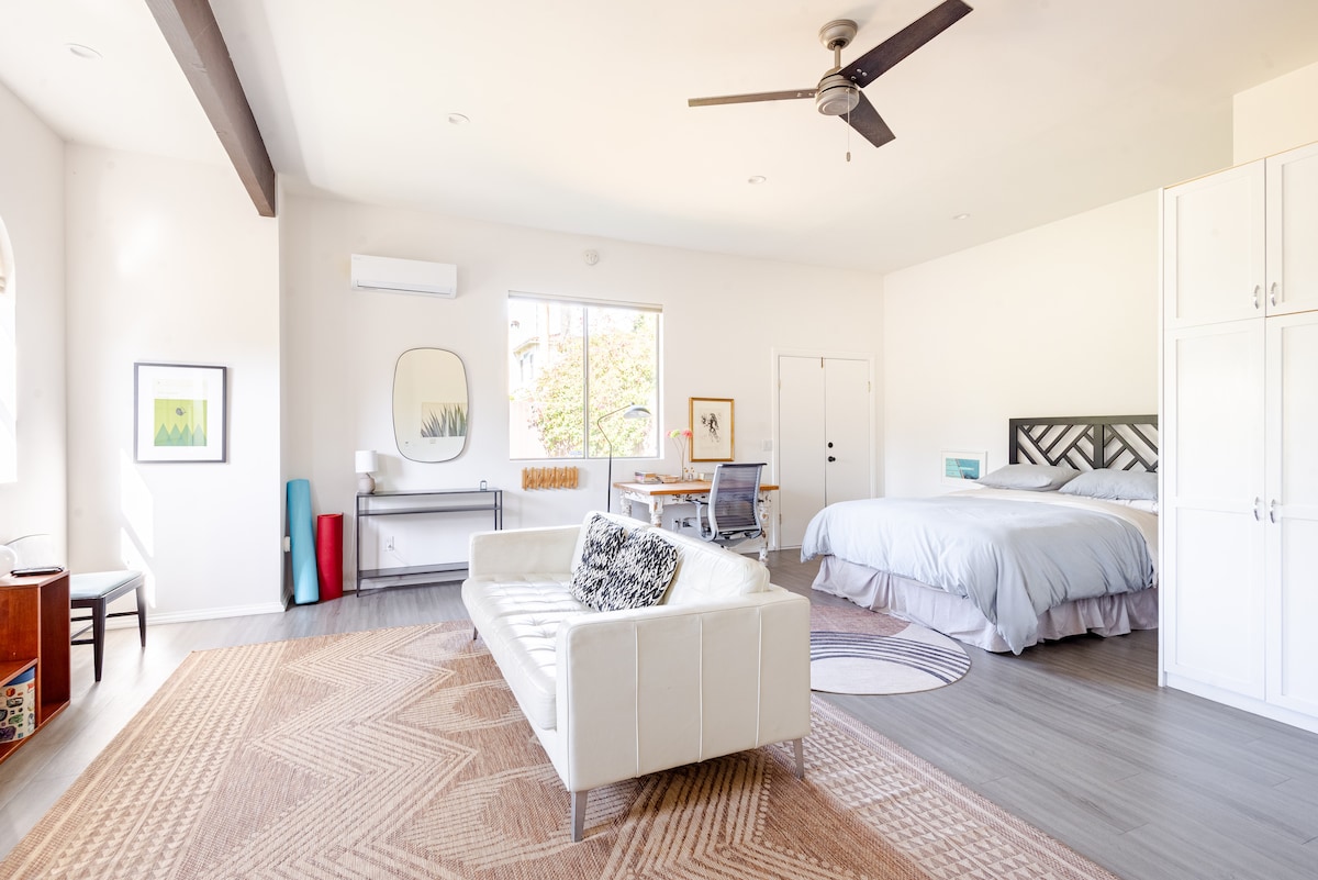 A bright and spacious studio is presented, featuring a California King bed with light bedding. A comfortable white couch is positioned on a large area rug, while a table and chairs are situated near the window, which offers natural light. Ample storage is provided by built-in cabinets.
