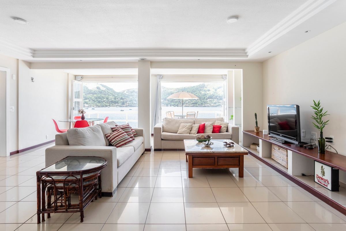 A light-filled living area features two sofas, a wooden coffee table, and a television unit. Large sliding glass doors open to reveal a patio with outdoor seating overlooking the sea. Natural light enhances the open and airy feeling of the space.