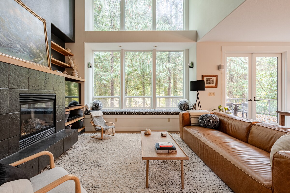 A spacious living area features a large leather sofa and a cozy armchair, inviting relaxation. A stone fireplace anchors the room, surrounded by shelves displaying decor. Tall windows allow natural light, with a custom window seat overlooking the forested views outside.