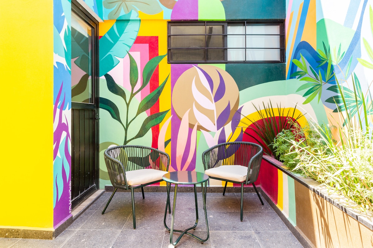 A vibrant terrace showcases a bold mural featuring lush tropical designs. Two black wicker chairs with light-colored cushions are positioned around a circular glass-top table. Greenery adds a natural element to the space, contributing to a lively outdoor ambiance.