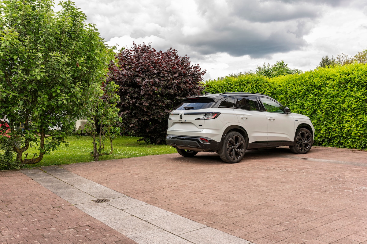 A parked white SUV is shown on a spacious paved area surrounded by lush greenery. Tall bushes and trees frame the space, providing a sense of privacy. The sky features soft, cloud-covered hues, contributing to a serene outdoor environment.