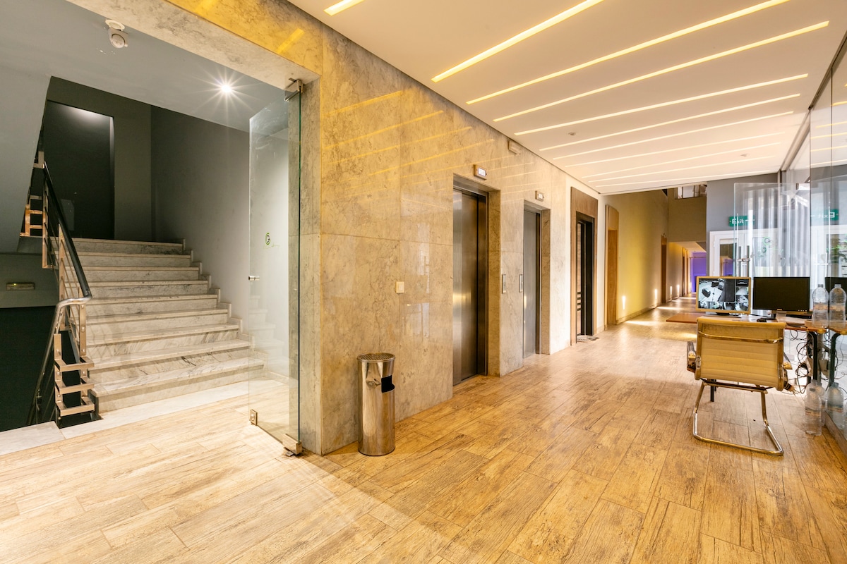 A well-lit and spacious lobby area is presented, featuring modern elevators and access to a staircase with sleek handrails. Natural wood tones and polished tile flooring contribute to a contemporary aesthetic. An area for reception or workspace is visible on the right.