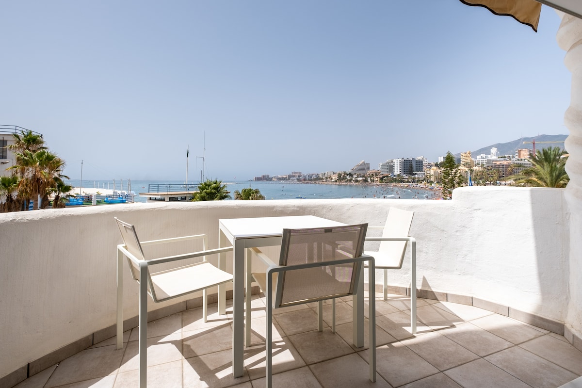A private terrace offers outdoor seating with a table and two chairs, overlooking the beach and marina. The horizon displays a clear sky, while nearby buildings and palm trees are visible, creating a relaxed ambiance.