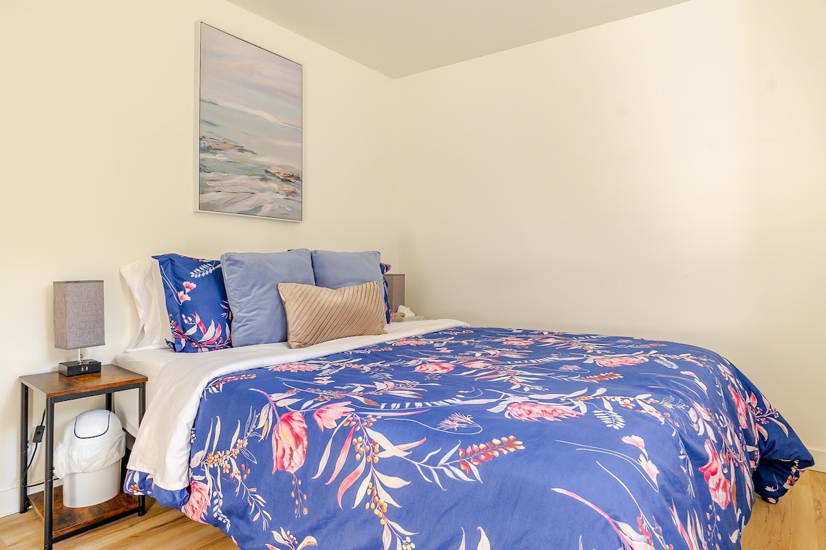 A spacious bedroom is adorned with a floral patterned bedspread in shades of blue and pink. A large painting of a serene coastal scene hangs above the bed. Two bedside tables with lamps are positioned on either side, contributing to the calming atmosphere.