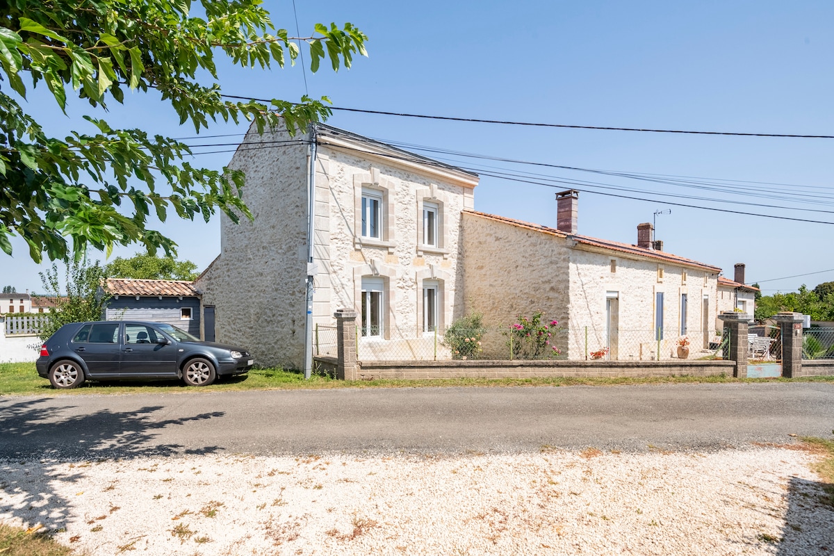 The exterior of a two-story stone house is shown, featuring light-colored walls and multiple windows. A car is parked along a gravel pathway. The surrounding area includes greenery and a clear blue sky, contributing to a serene setting.
