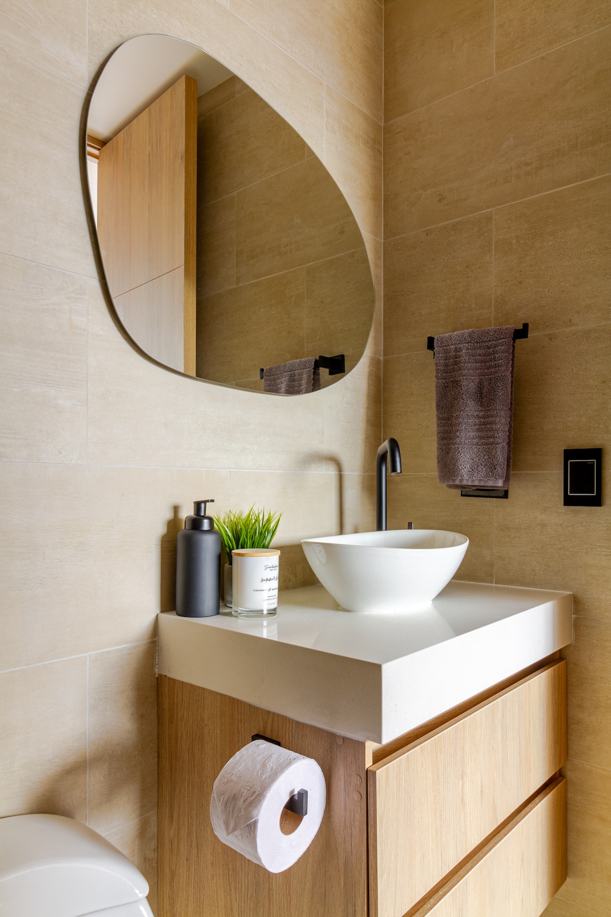 A modern bathroom features a light-colored wall with smooth tiles. A round mirror hangs above a minimalist vanity with a white basin and wood cabinetry. A sleek black faucet and a small plant add to the decor. A towel and toilet paper are neatly arranged within reach.