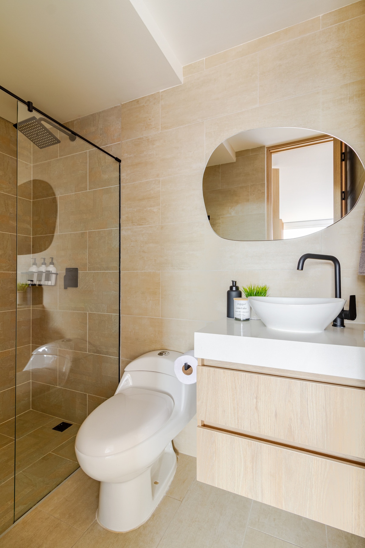 The bathroom features a modern design with beige tiled walls and flooring. A circular mirror is positioned above a sleek basin, while a glass shower enclosure is visible. A minimalist toilet is located beside a wooden vanity with storage below.