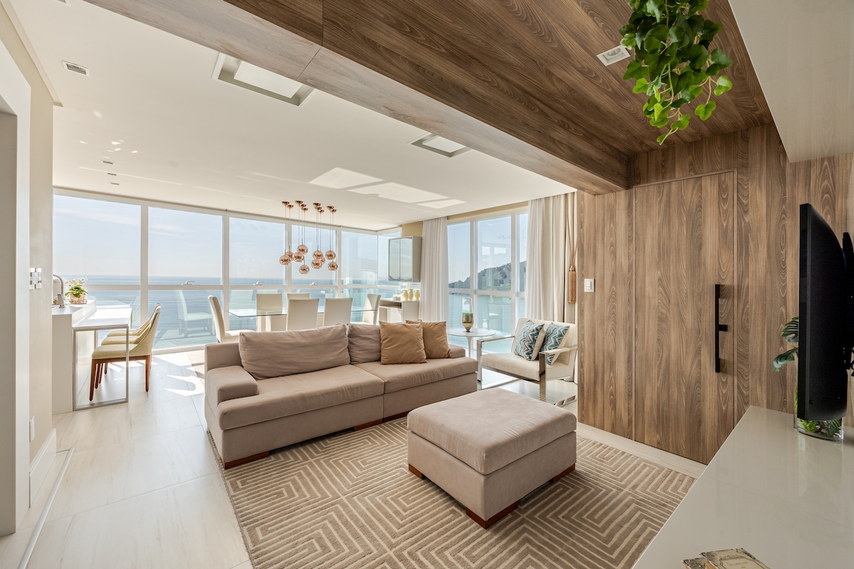 A modern living area features a spacious sectional sofa and an ottoman, positioned on a neutral-toned area rug. Large windows provide panoramic views of the ocean and let in ample natural light. A dining area with elegant pendant lights is visible in the background.
