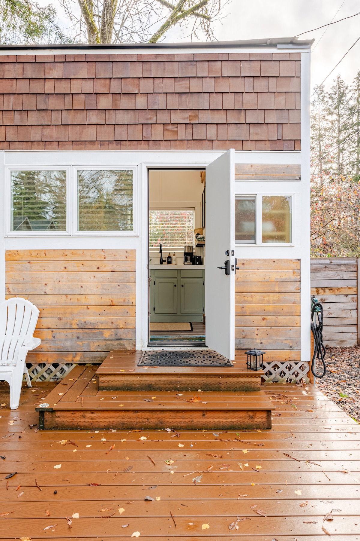 The entrance of the tiny home features a welcoming open door, revealing a glimpse of the kitchen inside. Steps made of wood lead up to the door, and a white chair sits nearby. A small lantern and scattered autumn leaves adorn the deck.