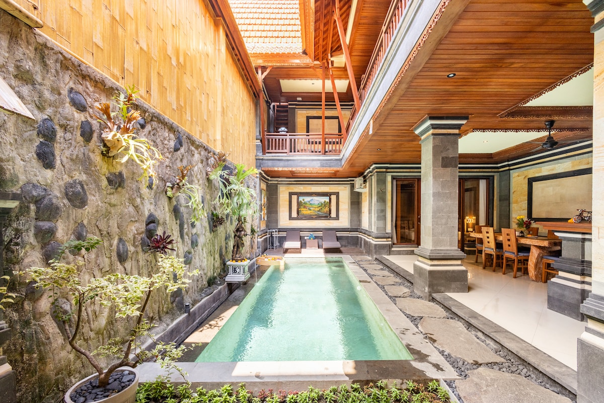 An inviting swimming pool is surrounded by a stone path and greenery. The villa features multi-level architecture with wooden details, large walls, and an open design, allowing natural light to fill the space. Dining and relaxation areas are visible in the background.
