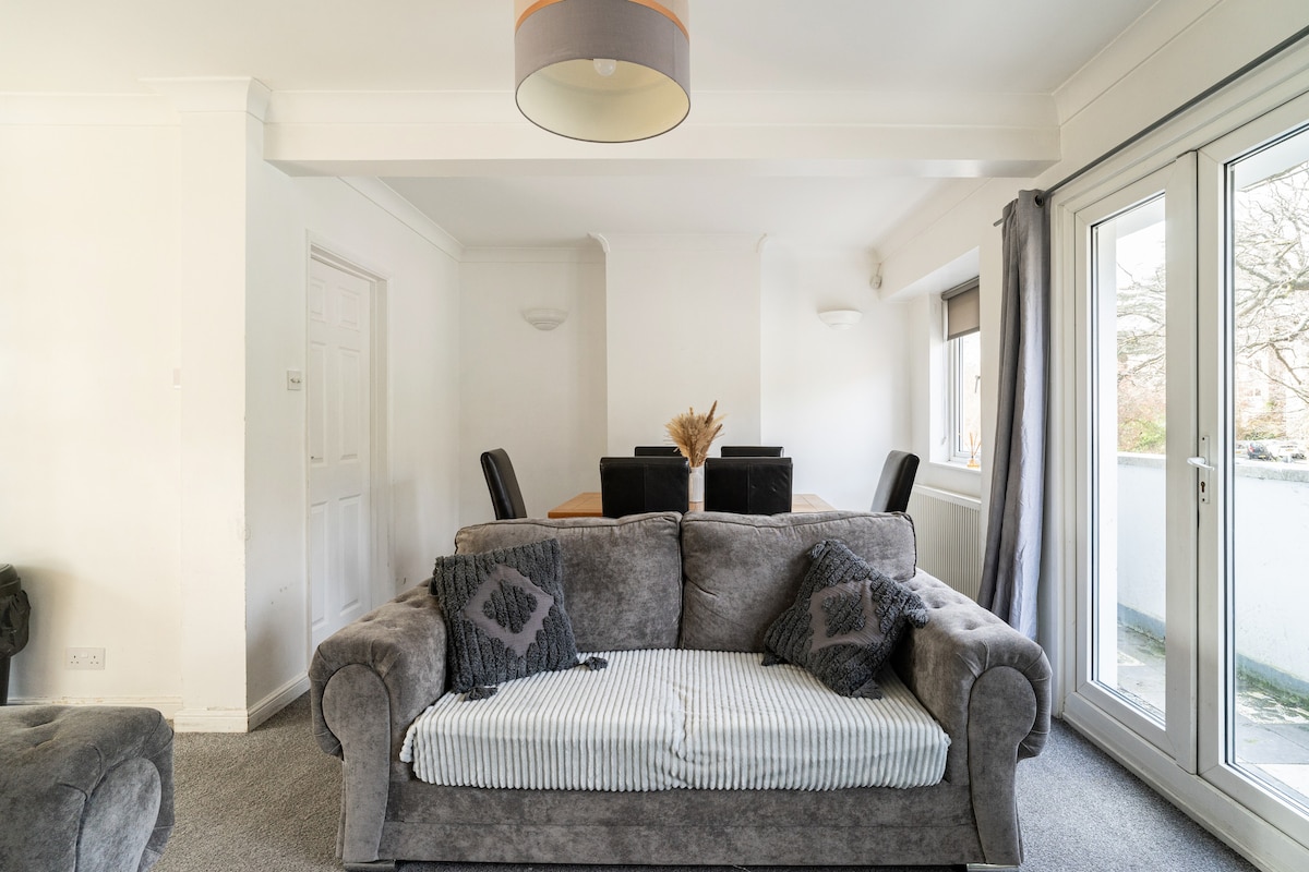 A comfortable living area features a plush gray sofa with decorative cushions. A neutral-colored rug covers the floor, while large glass doors provide natural light and views to the outside. A dining table with chairs is positioned in the background, emphasizing the open layout.