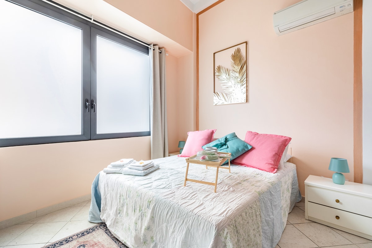 A light-filled bedroom features a spacious bed adorned with cozy bedding and accented by pink pillows. Two large windows provide ample natural light, and a bedside table holds a small tray. A modern air conditioning unit is mounted above the bed.