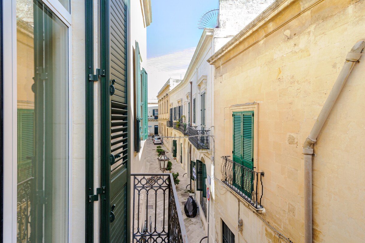 A view from the balcony showcases a narrow street lined with historic buildings featuring classic balconies and green shutters. The sunlight highlights the warm stone facades, while parked vehicles and potted plants can be seen along the cobblestone path.