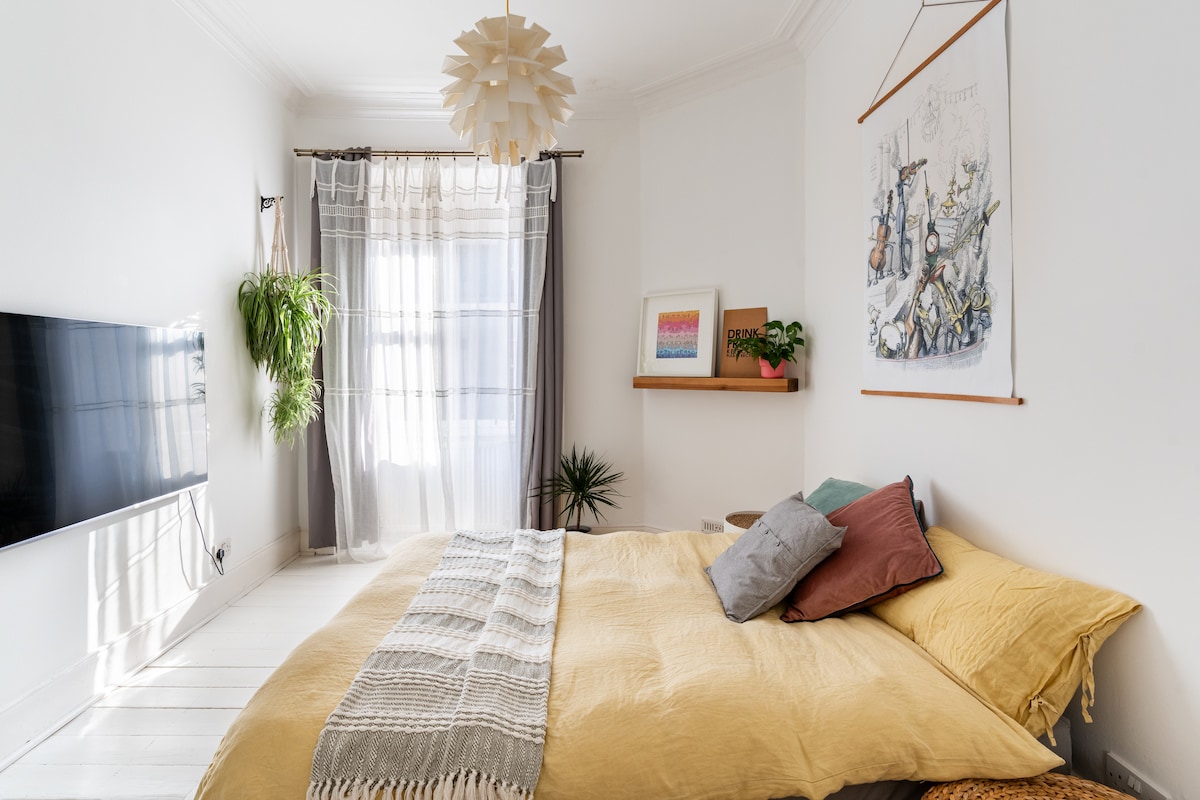 A bright bedroom features a spacious bed dressed in soft yellow linen, accented by a variety of decorative pillows. A light fixture hangs overhead, while a large window allows ample natural light to fill the room. Green plants add a touch of nature beside a wall-mounted shelf.