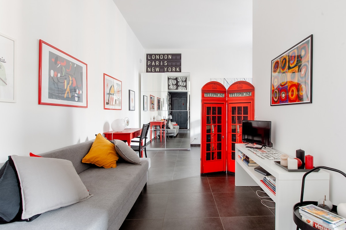 A modern living area features a gray sofa adorned with colorful cushions. Bright red accents, including iconic phone booths, contrast with the white walls. A dining area is visible in the background, decorated with framed art and natural lighting enhances the space.