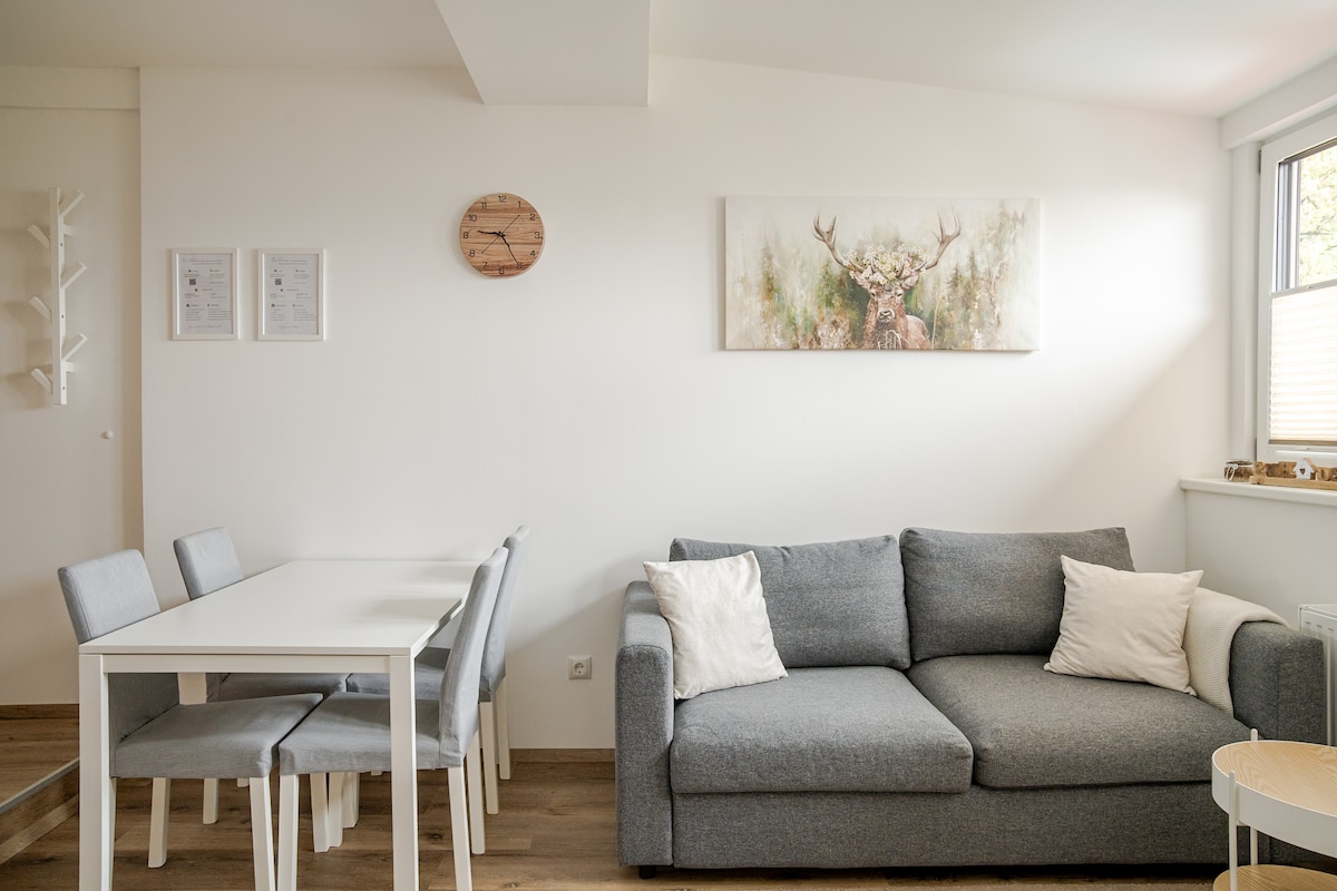 A light-filled living area features a gray sofa complemented by two decorative pillows. A white dining table is surrounded by four matching chairs. A wall-mounted clock and framed artwork add character to the space, with natural light streaming through the nearby window.