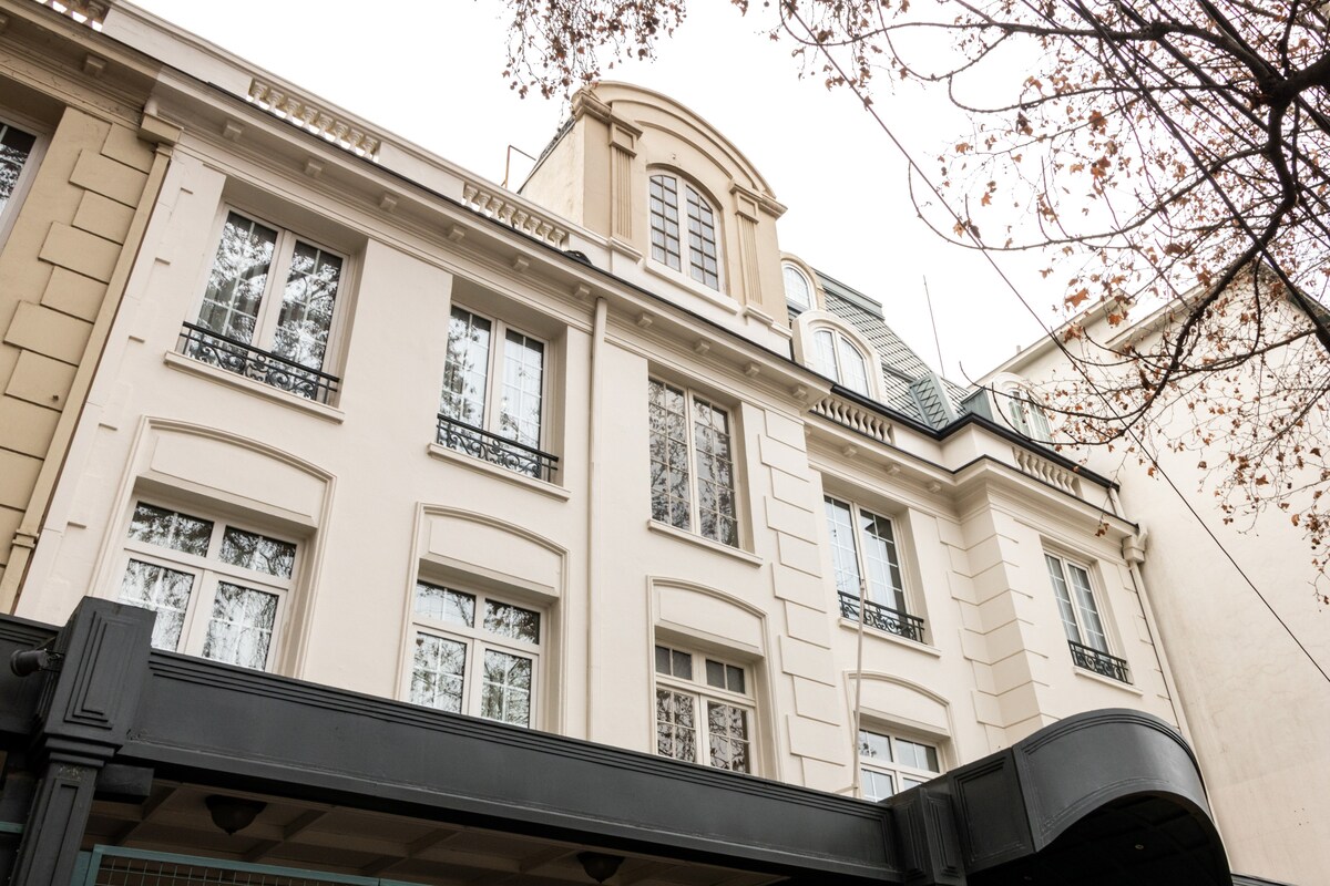The exterior of a classic French-style building is depicted, featuring elegant architectural details. Large windows are framed by delicate wrought iron balconies. A welcoming entrance is highlighted by a dark awning, while tree branches with sparse leaves provide a natural canopy above.