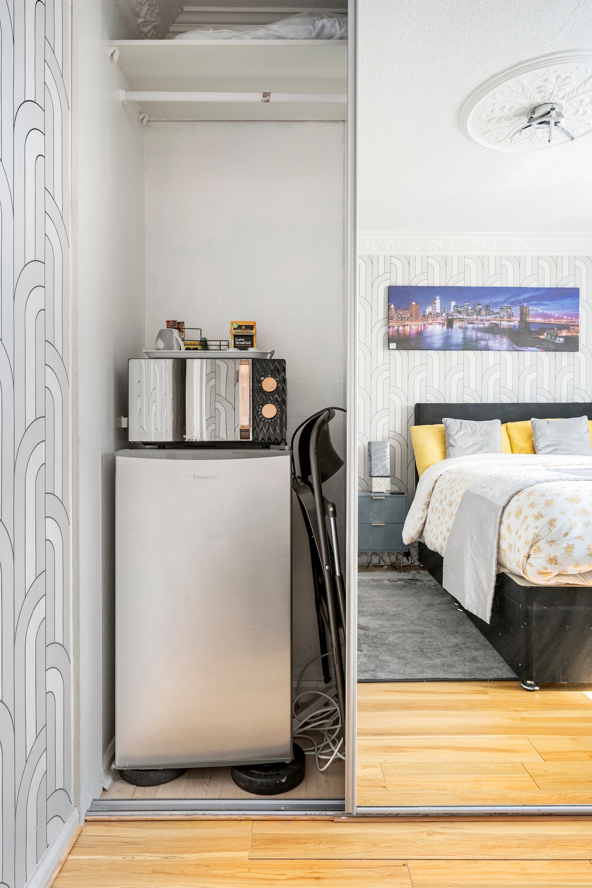 A compact corner features a silver refrigerator and a black microwave, both available for guest use. The mirrored wardrobe door reflects the cozy sleeping area with a bed dressed in light-colored linens and accented by decorative pillows.