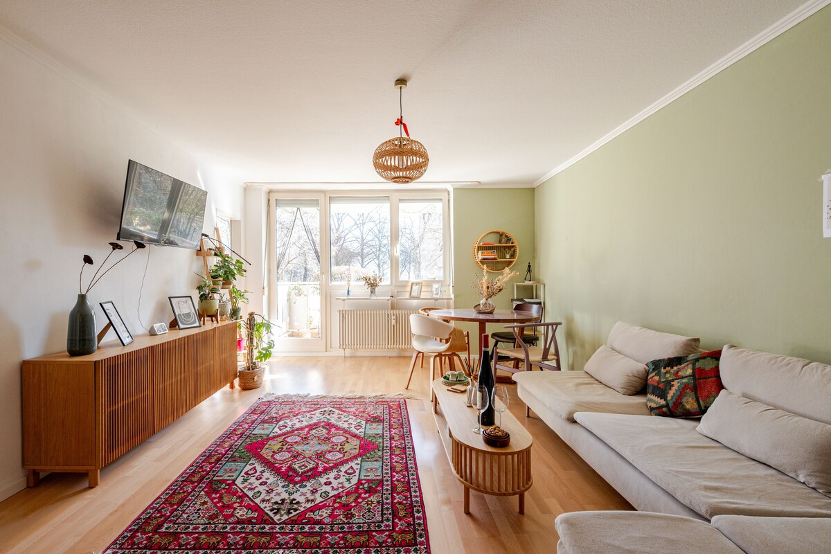 A bright living area features a light-colored sofa and a wooden coffee table. Colorful cushions are arranged on the sofa. A vibrant rug lies on the wooden floor, and large windows allow natural light to enter, providing views of the greenery outside.