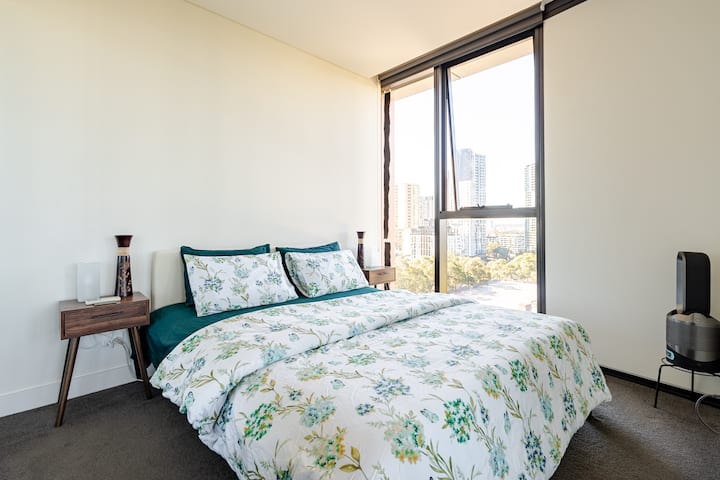 A bright bedroom with large floor-to-ceiling windows offering a city skyline view. The bed is dressed in floral-patterned bedding with green accents. Minimalist wooden bedside tables hold decorative items, and the room has a clean, modern feel