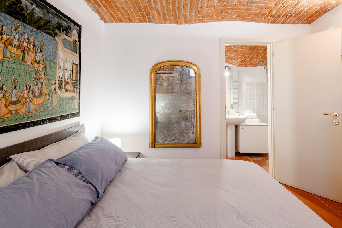 A cozy bedroom features a spacious bed dressed with soft linens, complemented by a vintage mirror above a small dresser. Exposed brick ceilings add character, while a door leads to an en-suite bathroom, designed with bright tiles and modern fixtures.