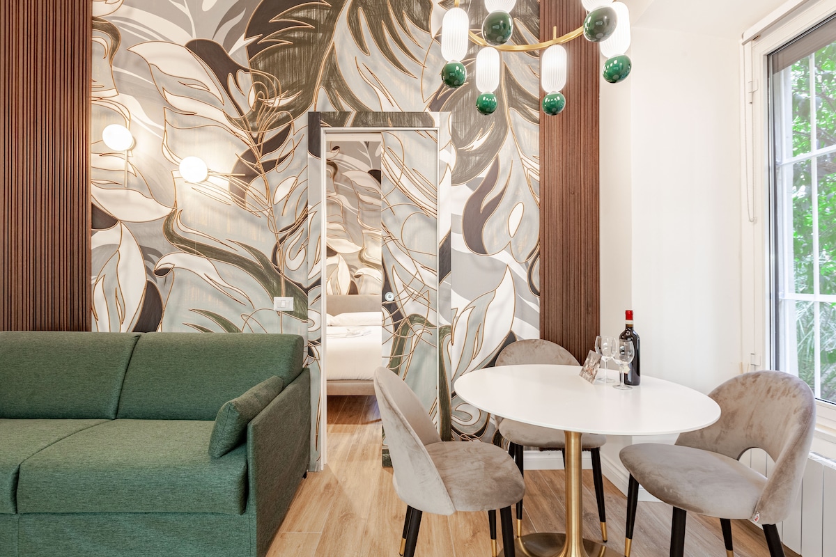 A stylish open space features a green sofa facing a round white table surrounded by plush chairs. The backdrop displays an artistic wallpaper with leafy patterns, and a modern light fixture hangs above. A door leads to a bedroom area, suggesting a cohesive design.