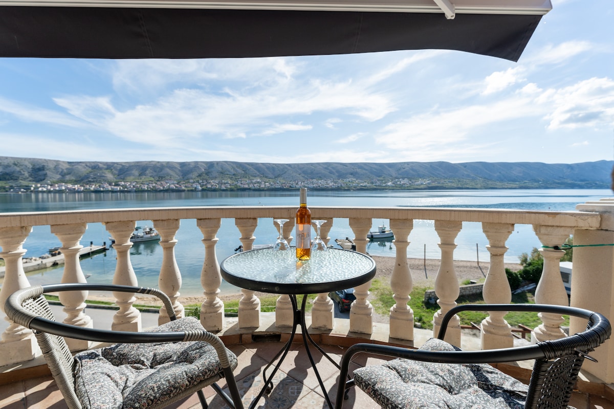 A balcony is shown overlooking calm waters, featuring a round glass-topped table and two woven chairs. An amber bottle is placed centrally on the table. The backdrop includes a view of rolling hills and distant boats along the shoreline, under a clear blue sky.