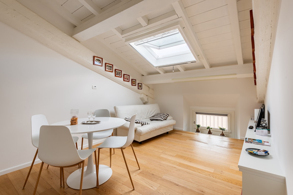 The open living area is illuminated by natural light from a skylight. A white round table is surrounded by four chairs, while a light-colored sofa is positioned nearby. Decorative items are displayed on a shelf, and potted plants are visible on the window sill.