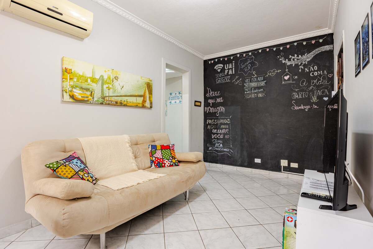 A living area features a light-colored sofa adorned with colorful decorative pillows. A large chalkboard wall displays various writings, while a flat-screen TV is positioned nearby. Natural light illuminates the space, enhanced by an air conditioning unit mounted on the wall.
