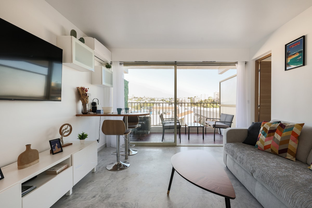 A modern living area features a comfortable sofa adorned with colorful cushions. A sleek coffee table sits in front, while a flat-screen television is mounted on the wall. Floor-to-ceiling glass doors provide access to a balcony with city views.