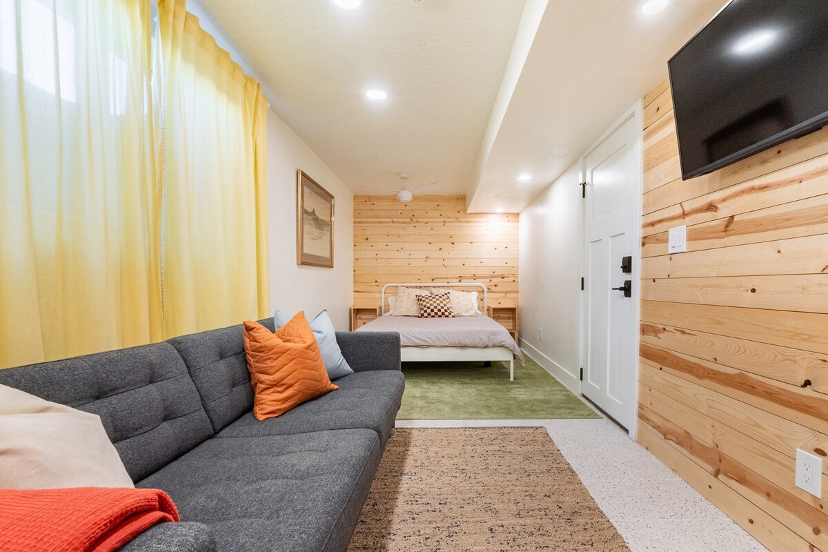 A cozy living area features a dark gray sectional sofa with colorful throw pillows, complemented by a textured area rug. A light wood-paneled wall serves as a backdrop for a queen-sized bed with simple bedding. Large windows dressed with sheer yellow curtains allow natural light to fill the space.